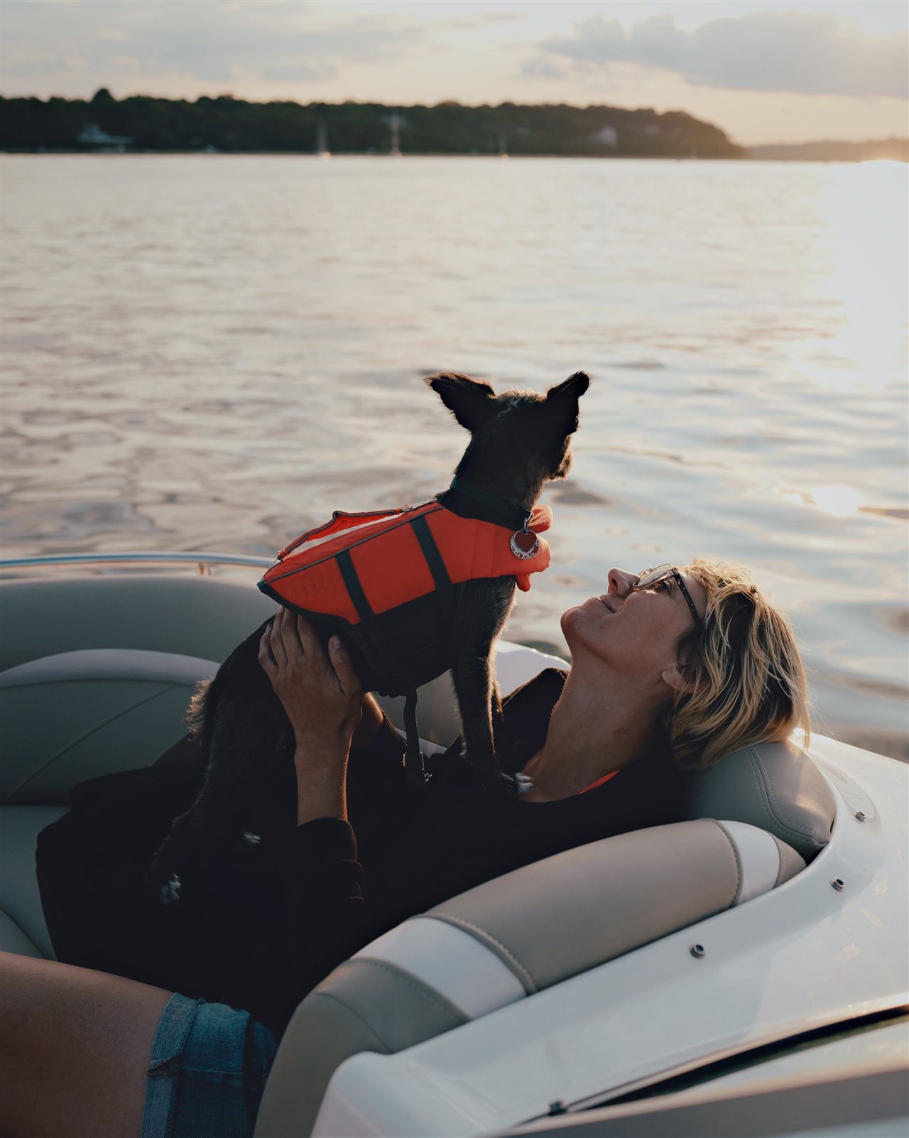 Woman on the back of a pontoon while her dog, wearing a life jacket, stands on her chest