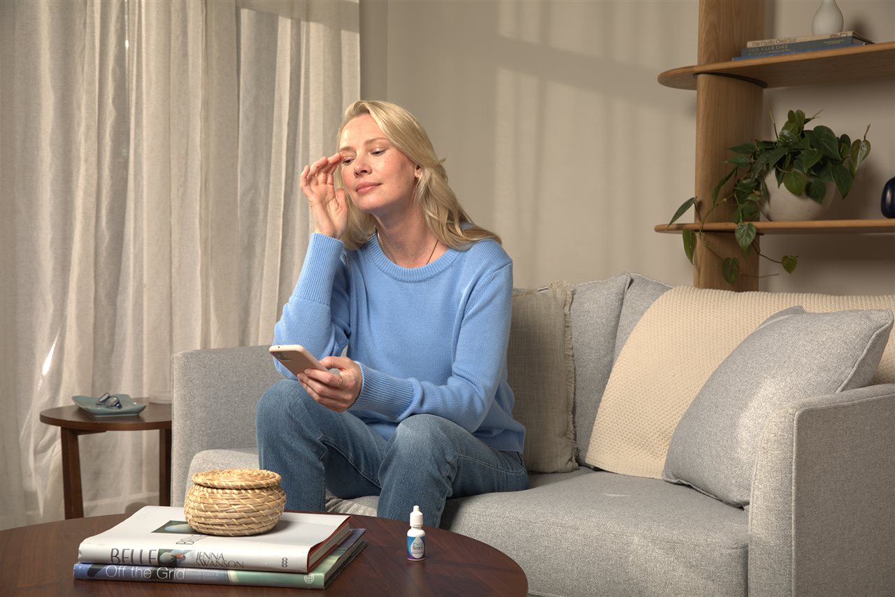 Woman sitting on the couch at home using her cell phone and looking at a bottle of eye drops on a near by coffee table.