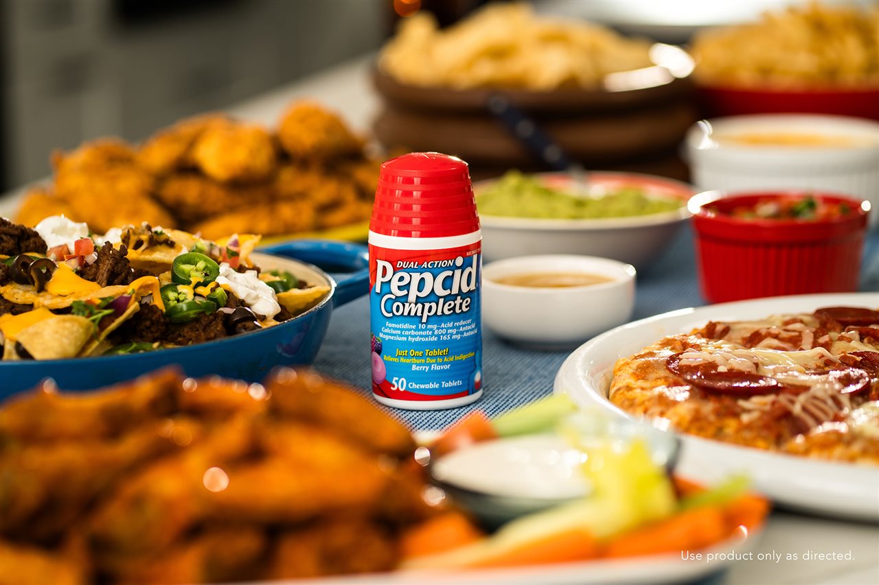 Pepcid on a table with dishes that may cause heartburn such as pizza, chicken wings, and nachos with peppers.