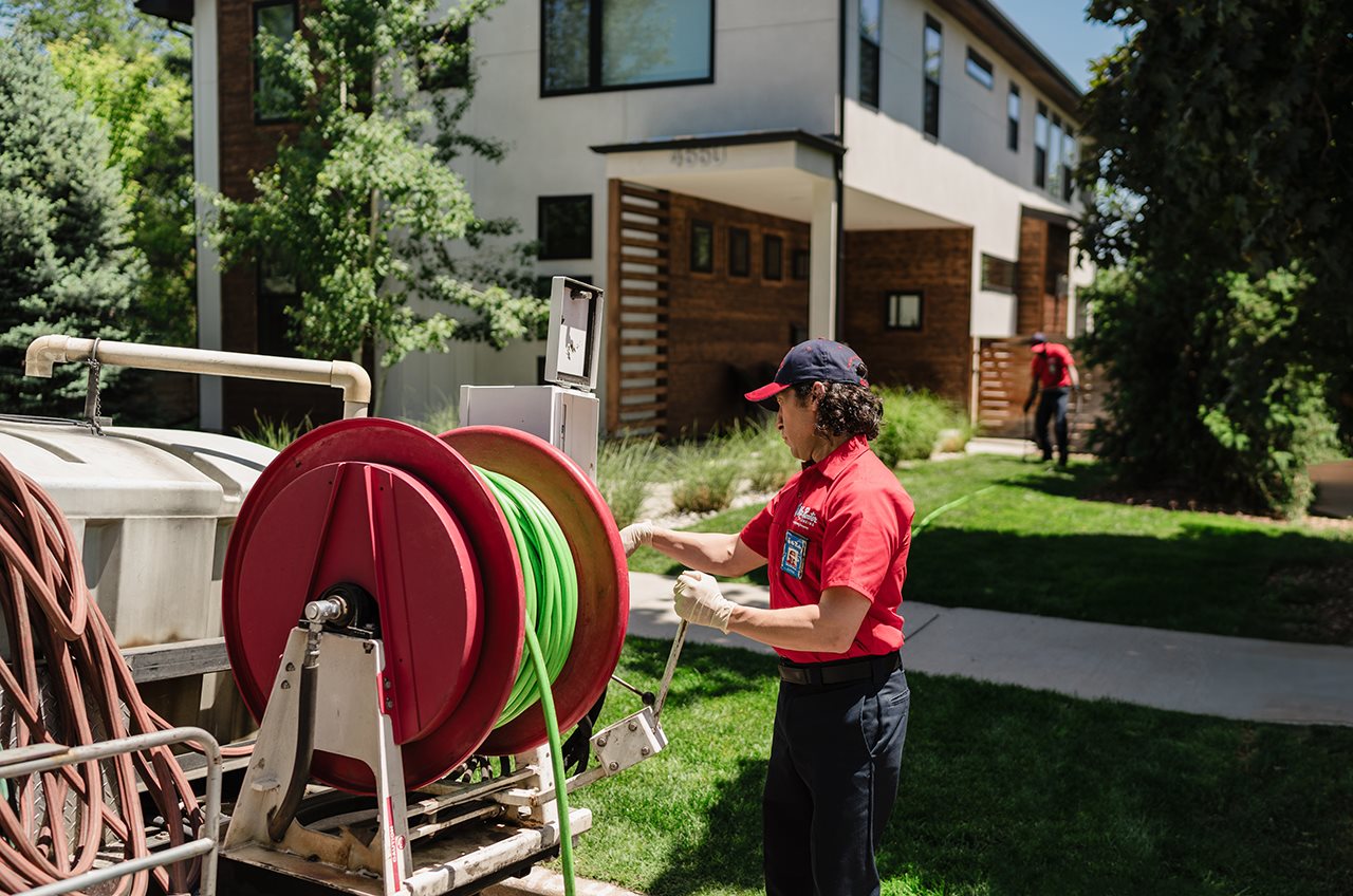 Female representative from Mr. Rooter pulling a hose off a reel in the back of a truck that connects to a device used to clean tree roots out of a pipe.