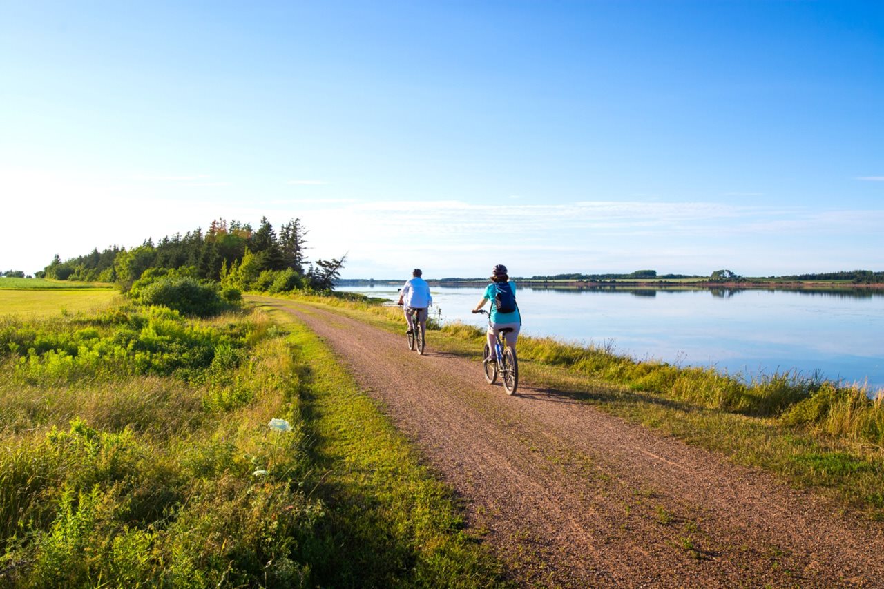 A couple ride bikes on a gravel road next to a body of water