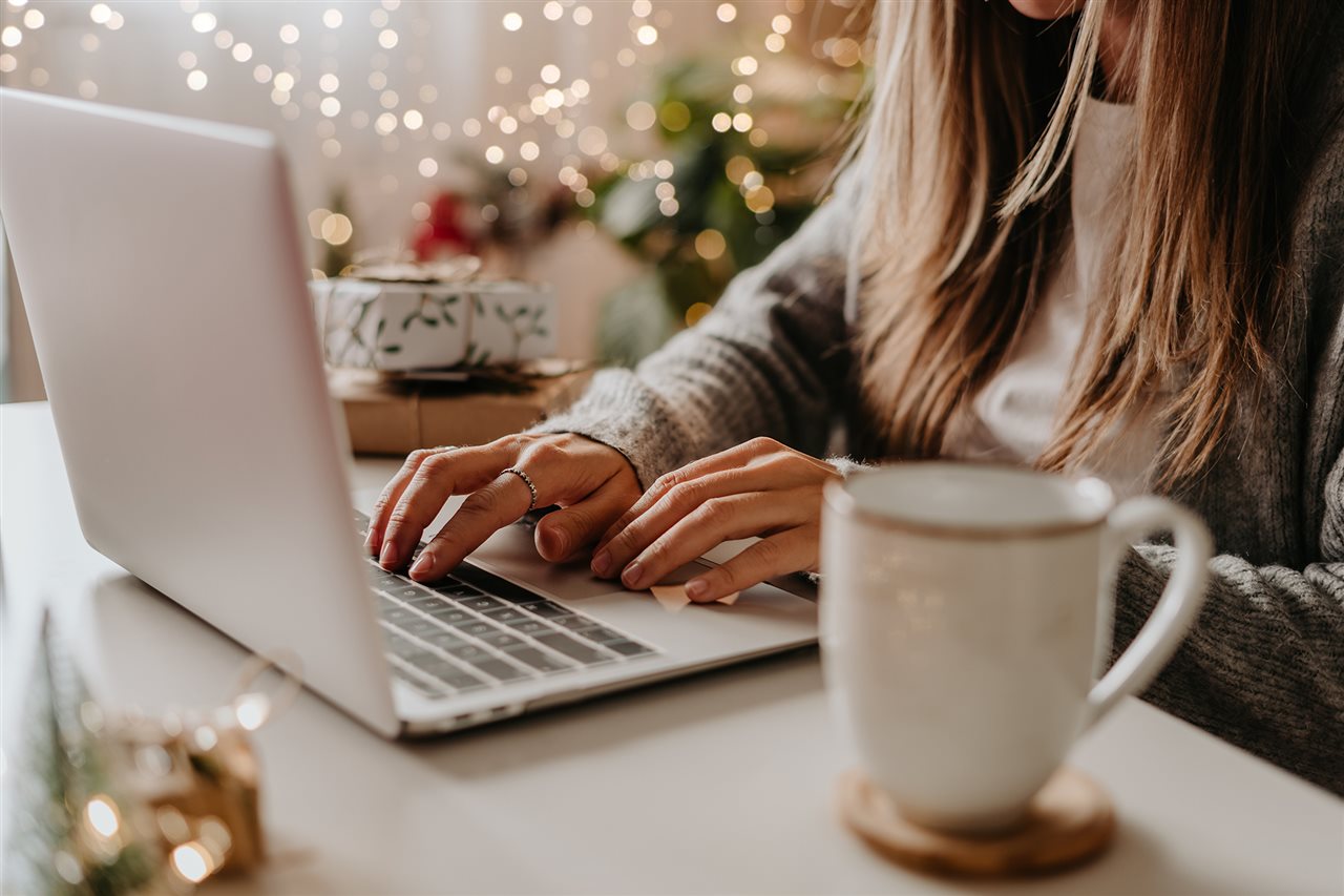 Close up of woman with her coffee on a laptop at home with white holiday lights in the background.