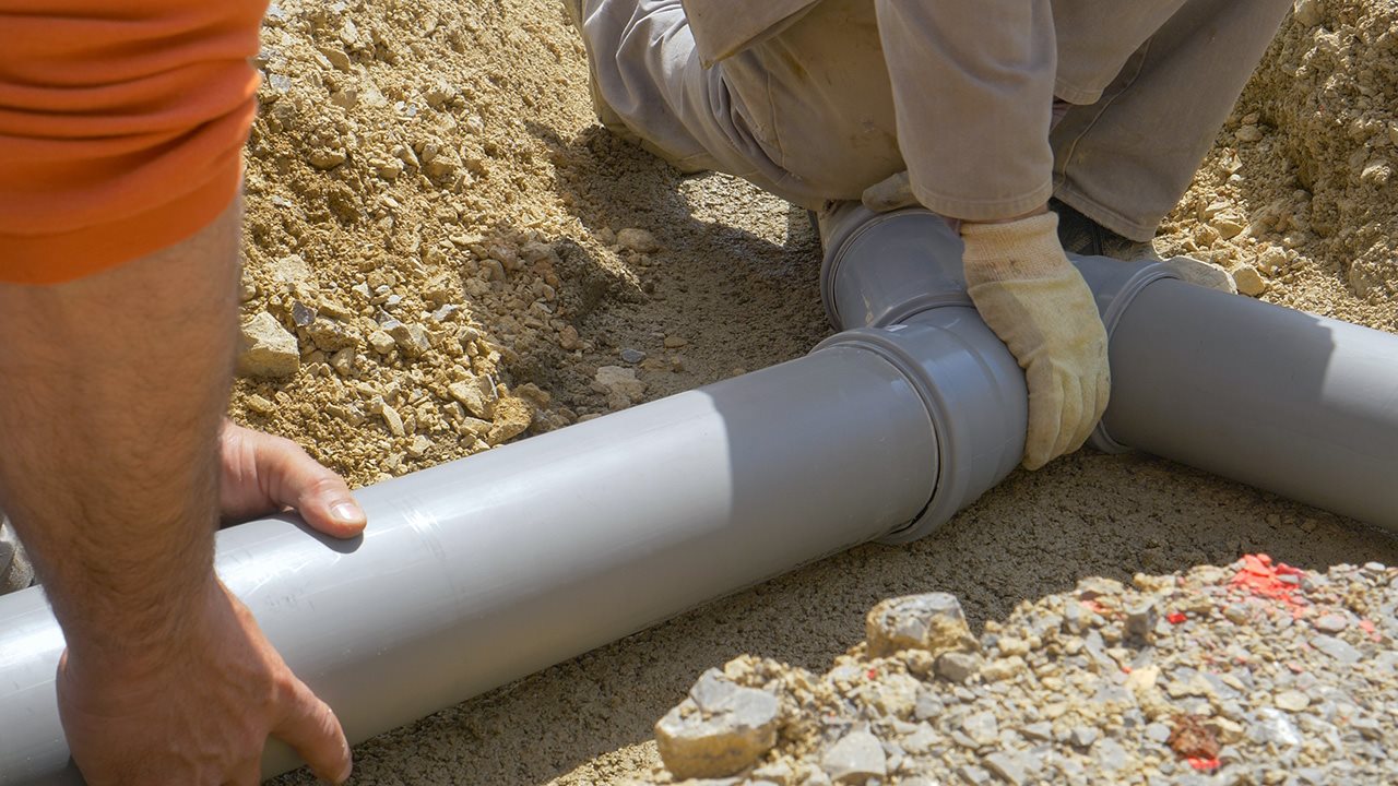 Two people hold together PVC pipes in the dirt.