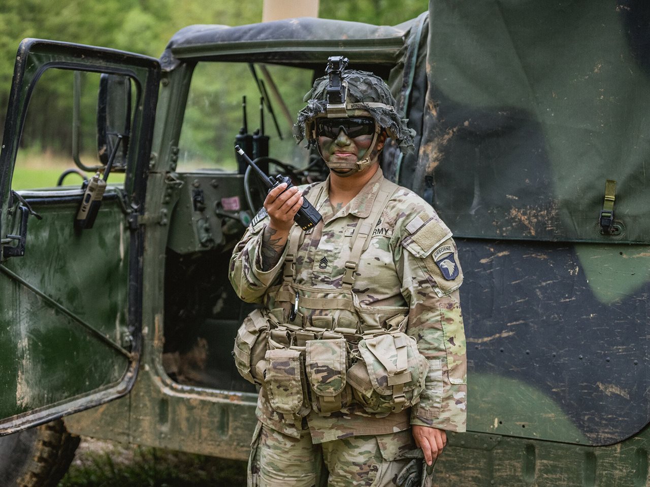 U.S. Army soldier dressed in fatigues talking on a walkie-talkie next to a truck.