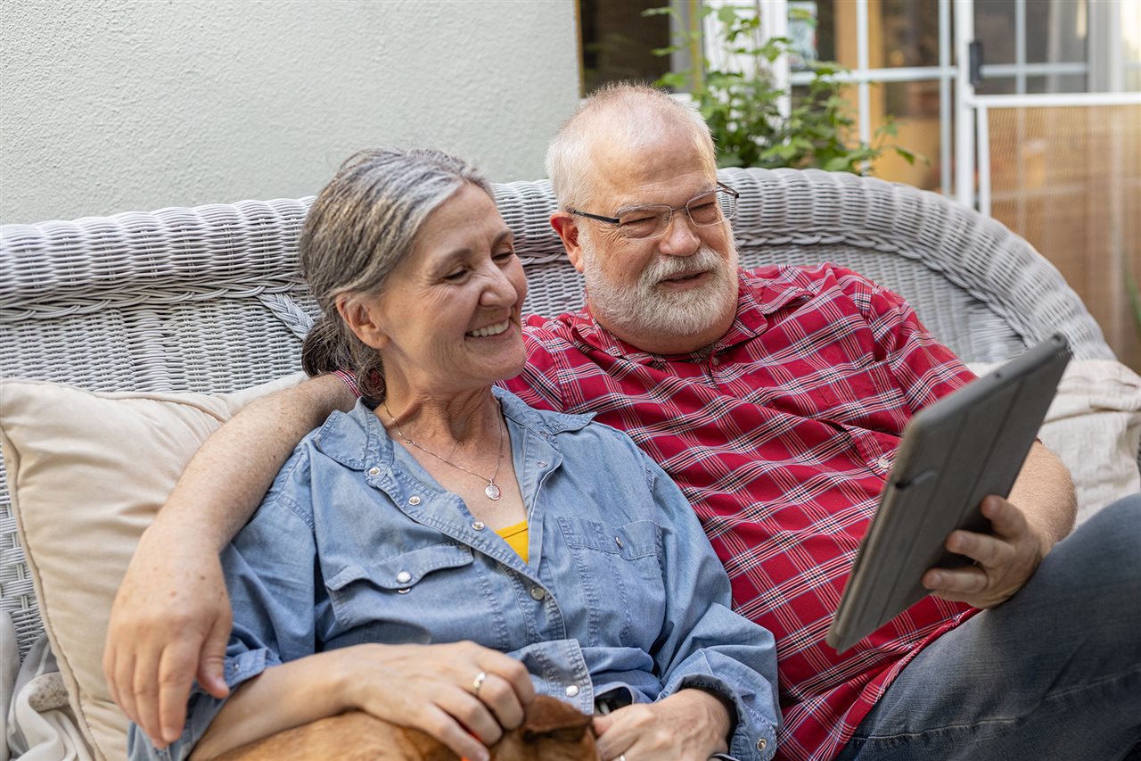 A senior couple looking over their options online using a tablet while sitting on wicker furniture outdoors.