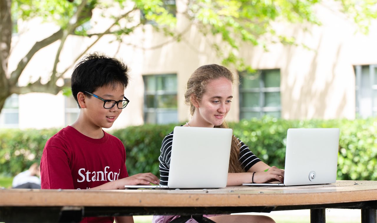 A male and a female student of Standford Online High School working on their laptops outside at a table.