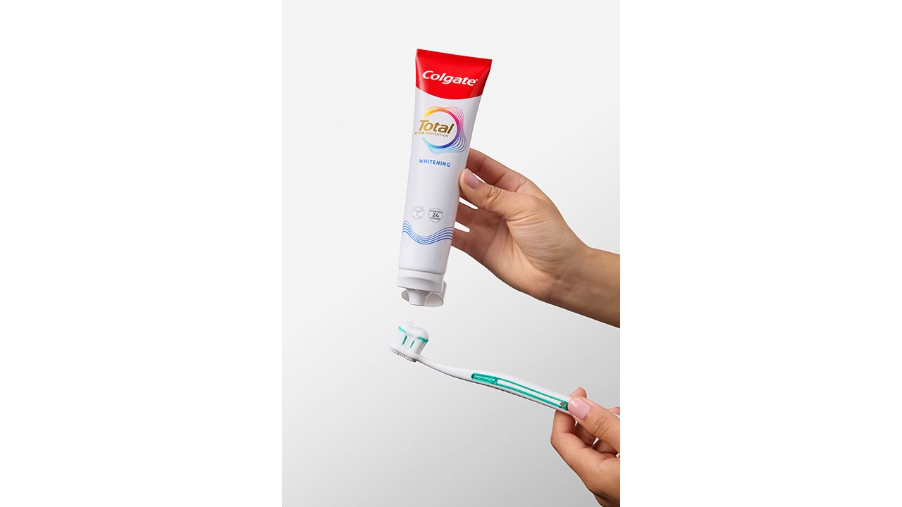A bottle of Colgate Total toothpaste is dispensed onto a toothbrush against a white backdrop.