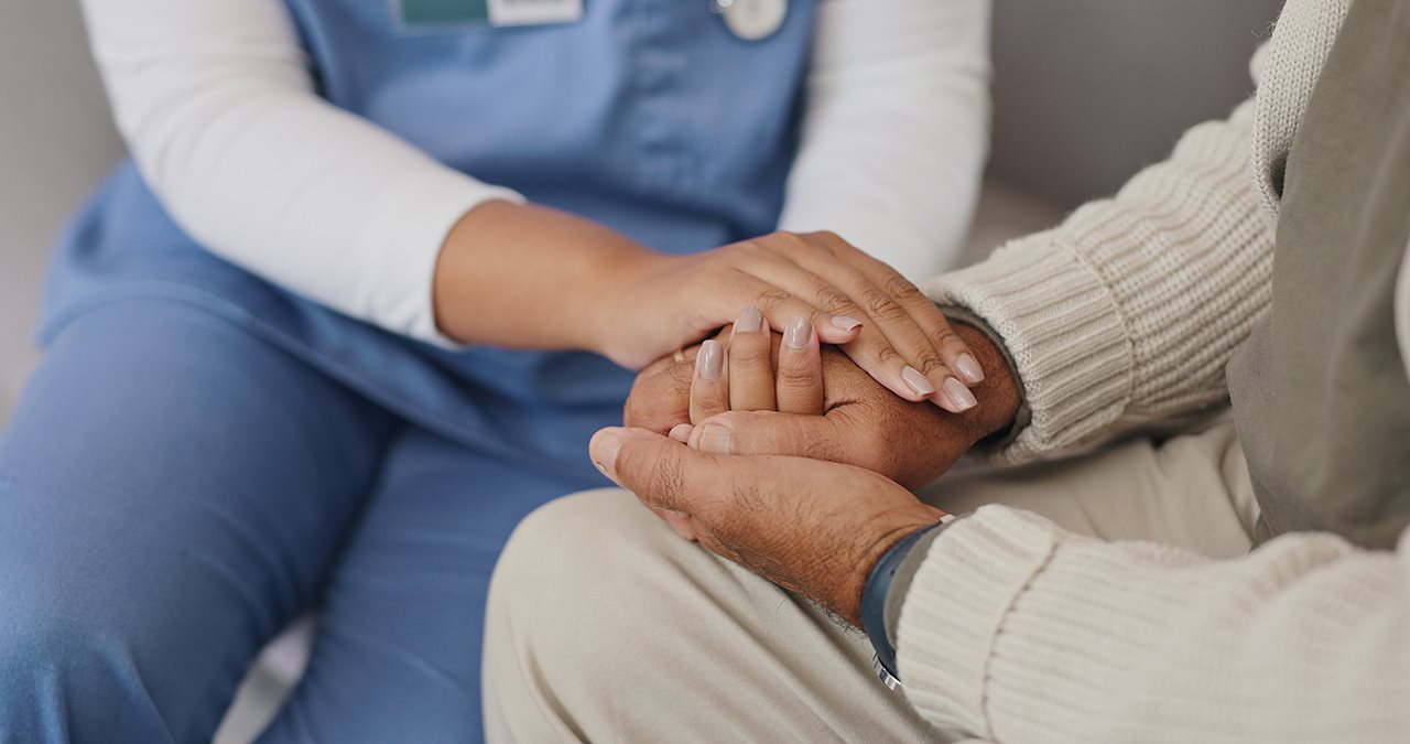 A female nurse holds hands with a patient in an off-white sweater.
