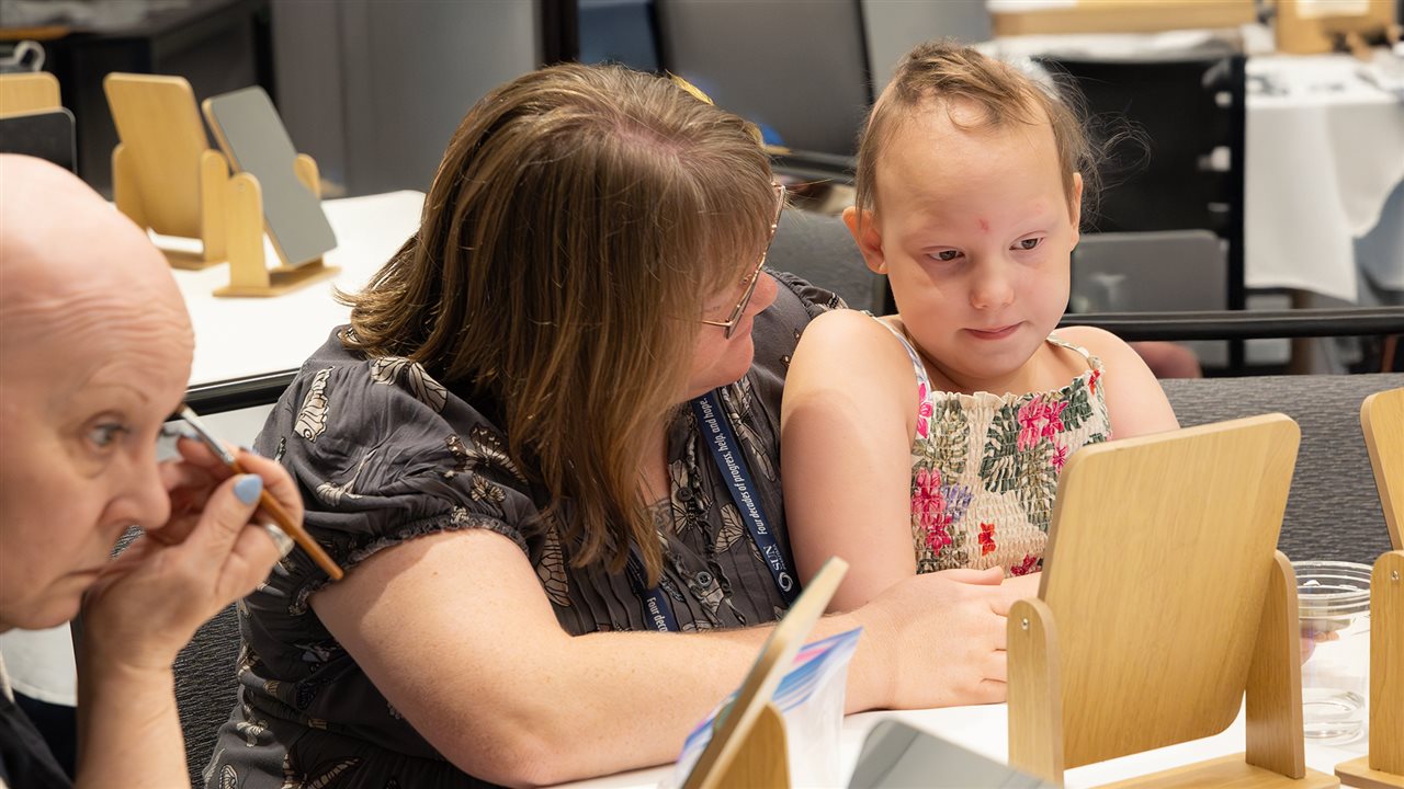 Little girl sitting at a table with a woman. Learn more at naaf.org/beyond-the-mirror