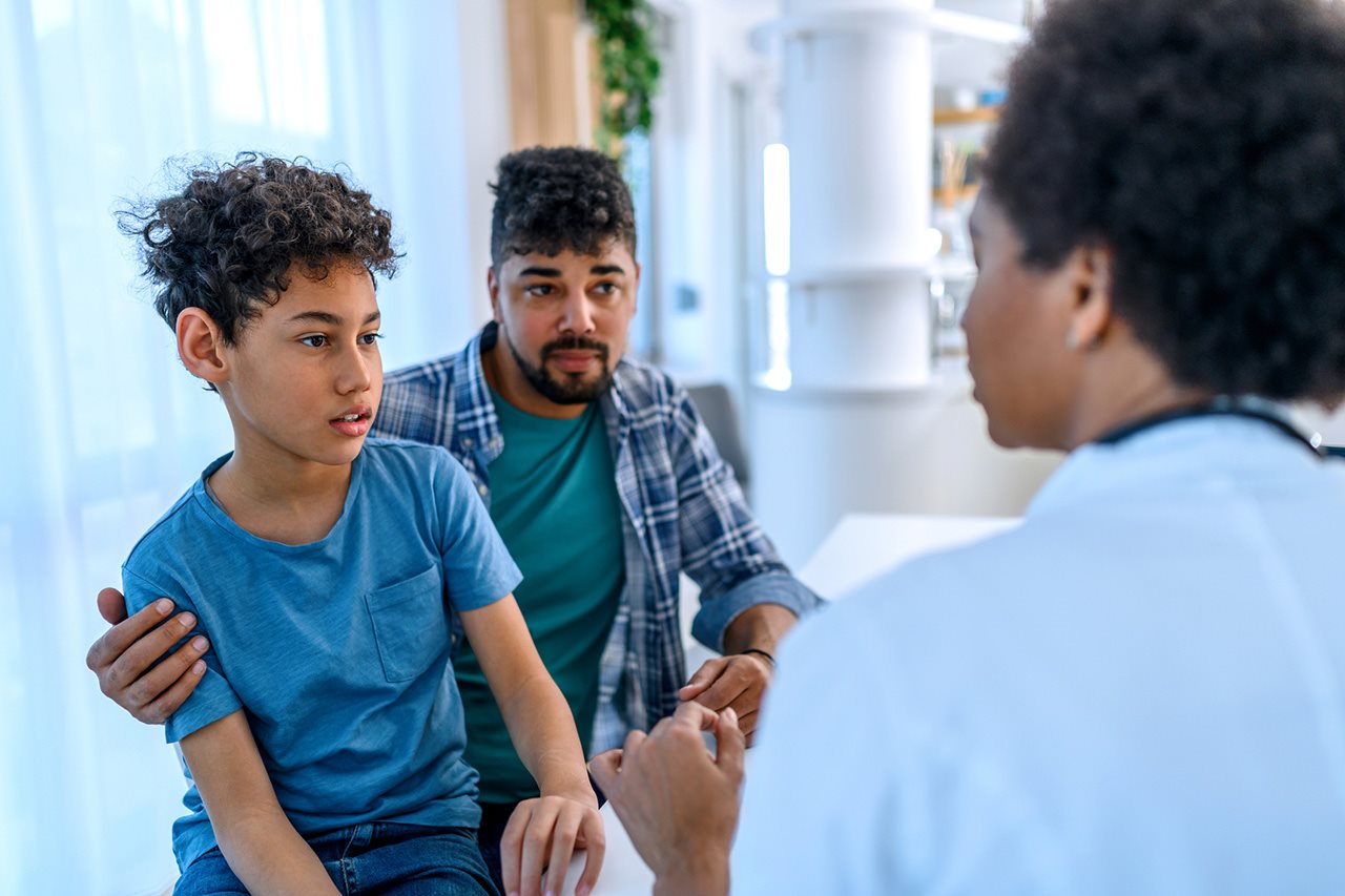 A father and son sit and listen to a doctor in her office.