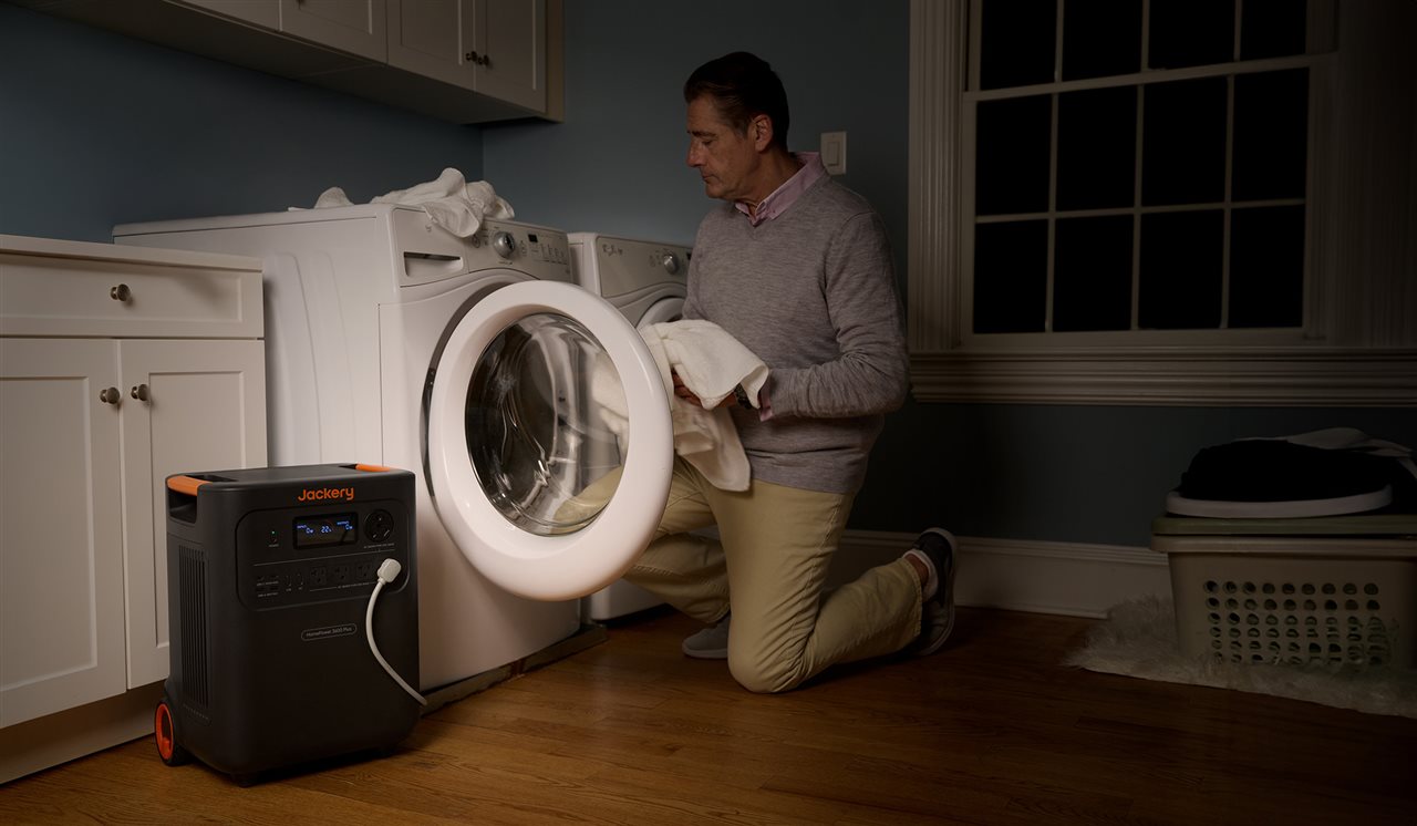 A man loads his washing machine in a dark laundry room, connected to a Jackery device.