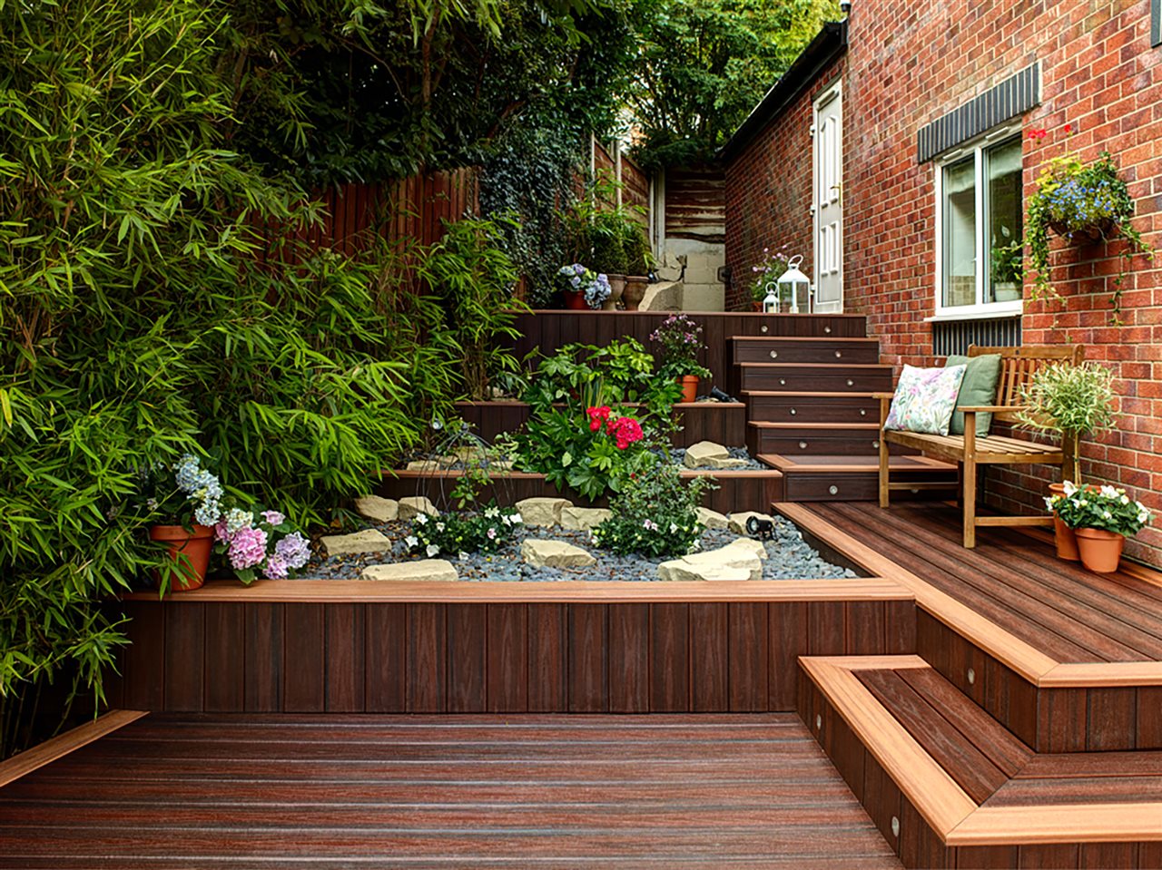 A lush wooden patio with stairs leading up to a brick house, flanked with a rock garden and plants.