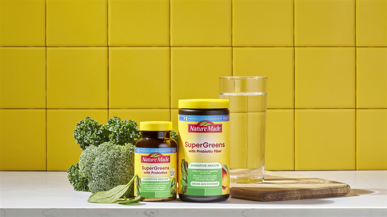 Bottle of Nature Made Supergreens with probiotics and a bottle of Nature Made Supergreen probiotic fiber on a counter top with vegetables.