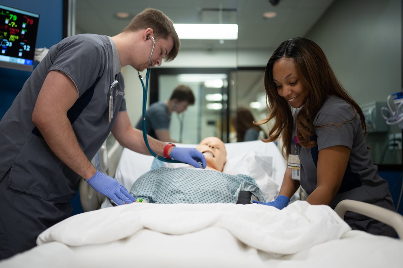 Two student practicing with a mock patient in a hospital setting.
