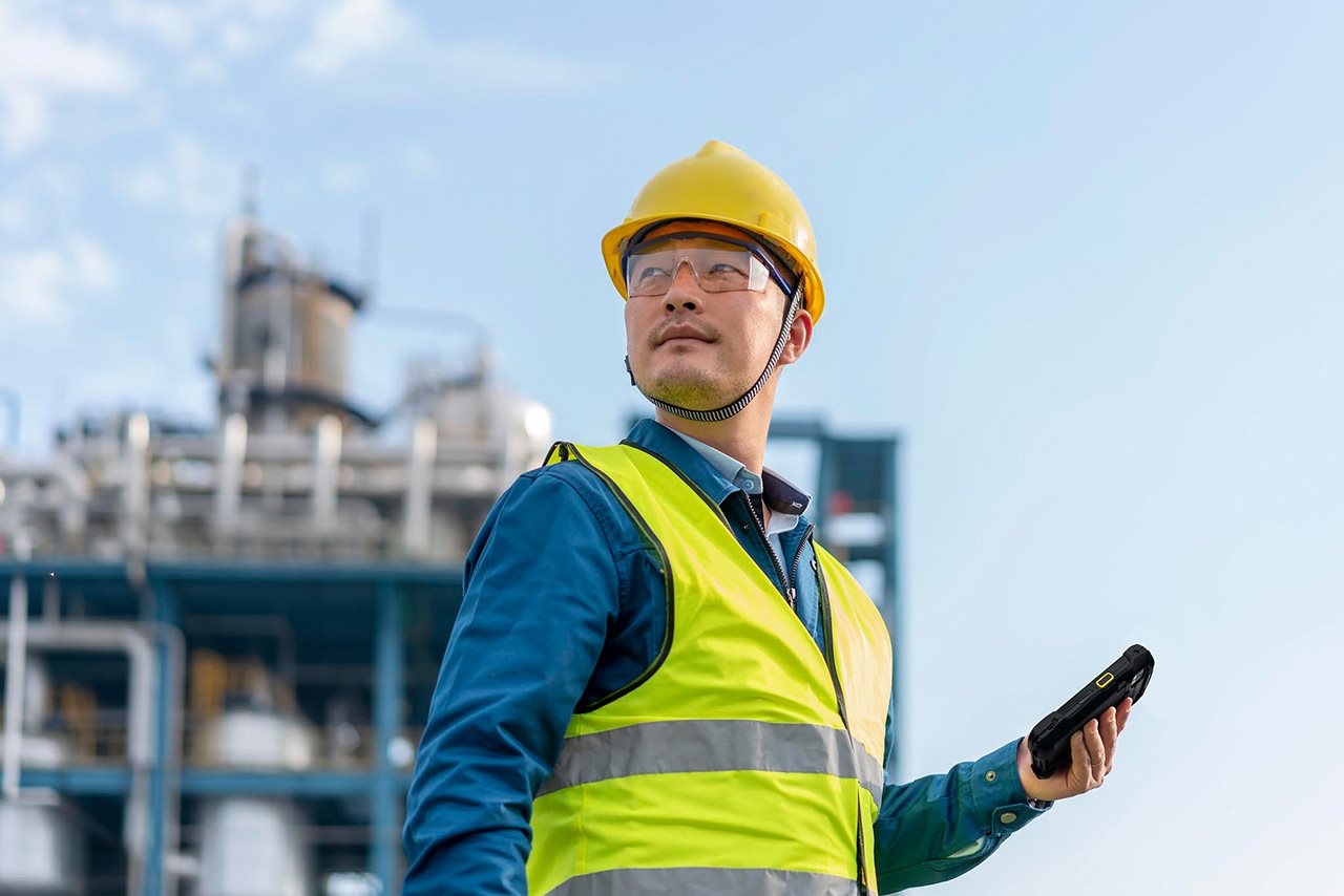 A man holding a mobile device stands in front of an industrial facility while wearing goggles and a helmet.
