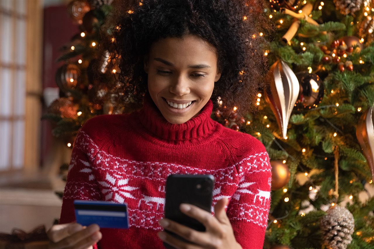 Smiling woman sitting in from of a Christmas tree shopping online with her credit car and her phone