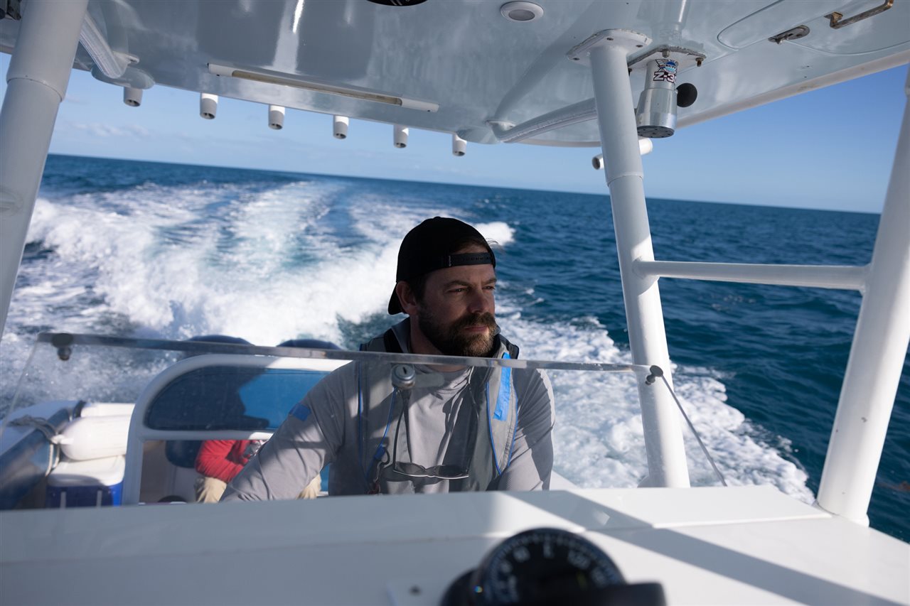Man, wearing a cap backwards, driving a boat across a lake