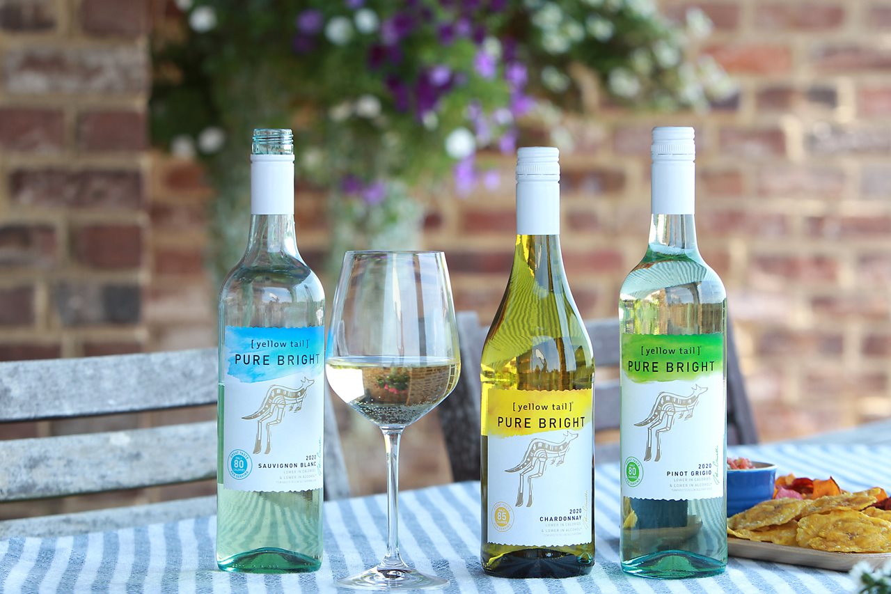 An assortment of [yellow tail] white wine in bottles, wine in a glass and a plate with food on checked table cloth.