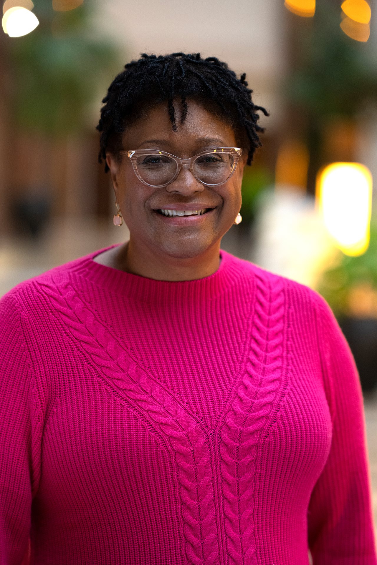 Smiling Lisa, wearing glasses and a pink sweater, standing outside.