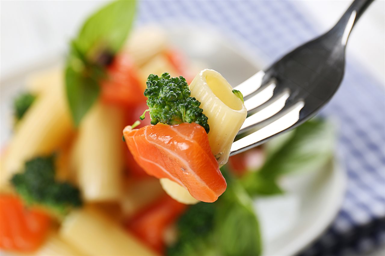 Close-up of a fork taking a piece of pasta, broccoli and salmon from a bowl.