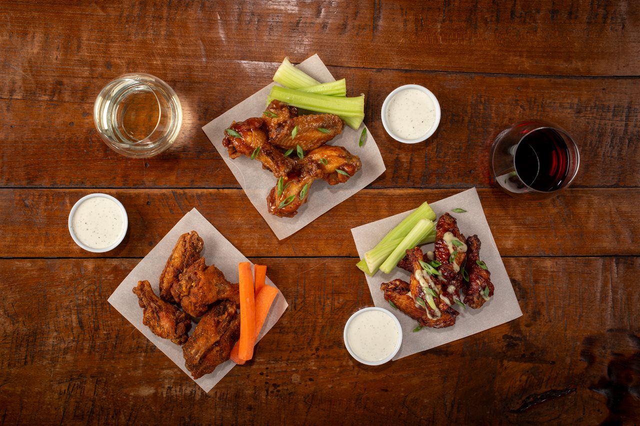 Small white plates with three varieties of chicken wings with dip, celery, and glasses of wine.