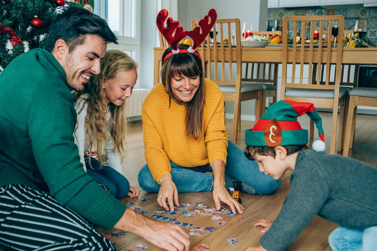 Family dressed in casual holiday clothing and wearing reindeer ears and elf hats while putting together a puzzle on the floor.