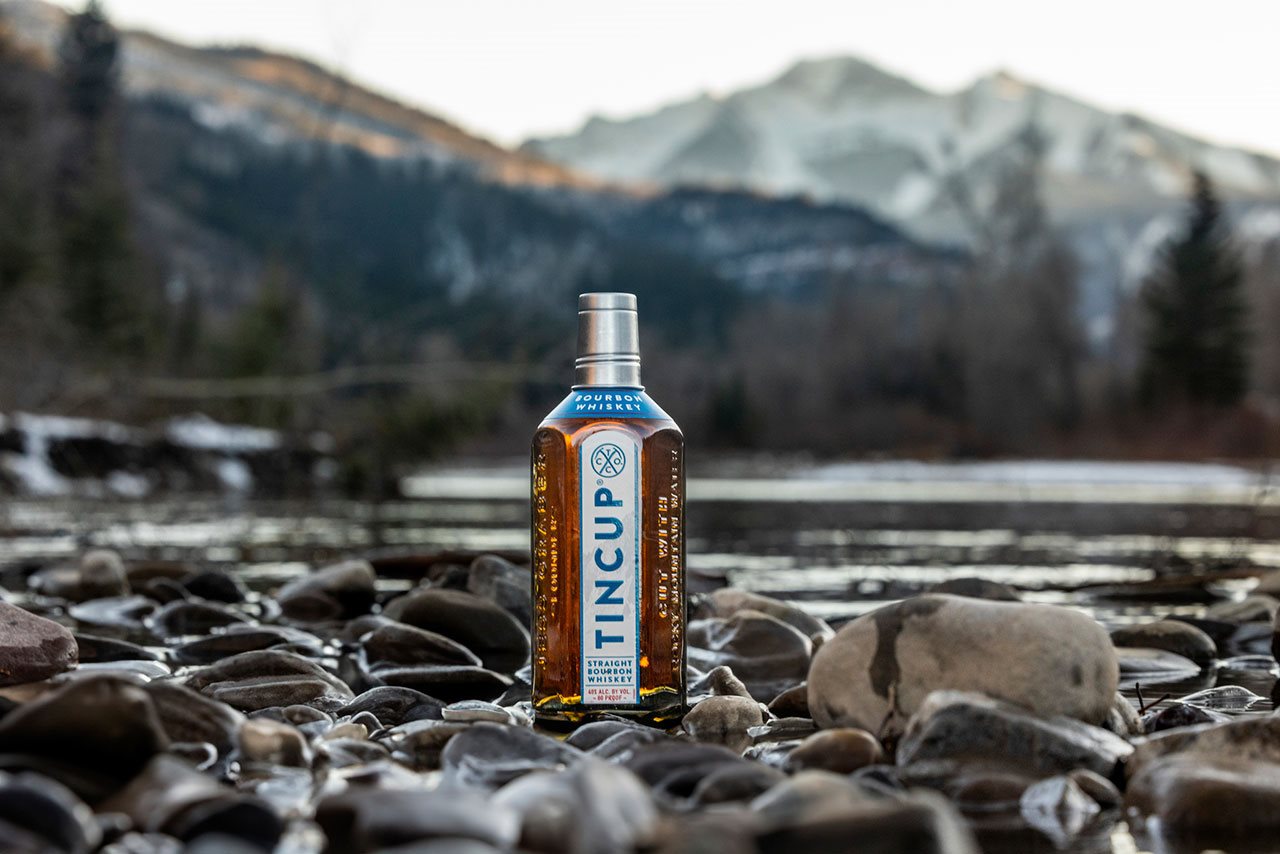 A bottle of Tincup whiskey outdoors sitting on a bed of rocks with a mountain in the background.