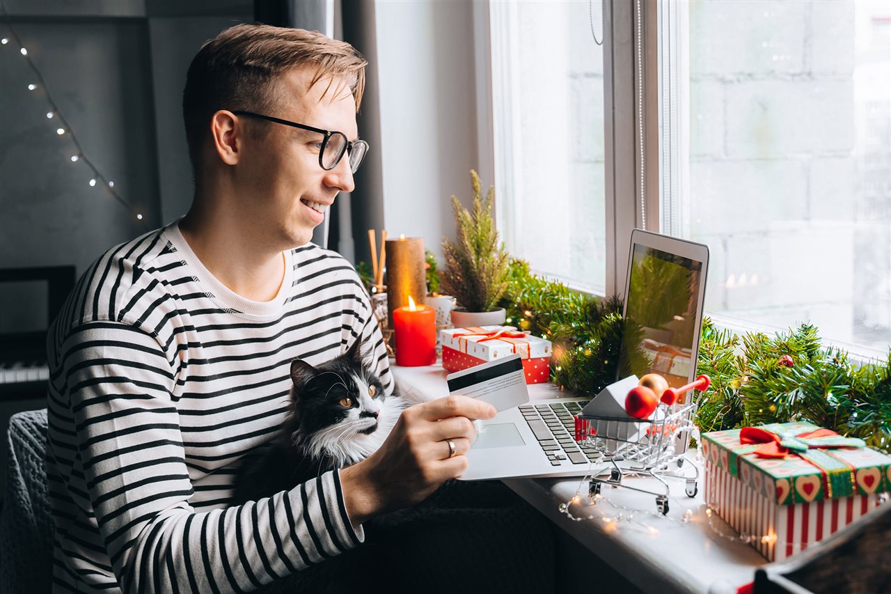 Man in striped shirt holiday shopping on his laptop while his can sits in his lap.