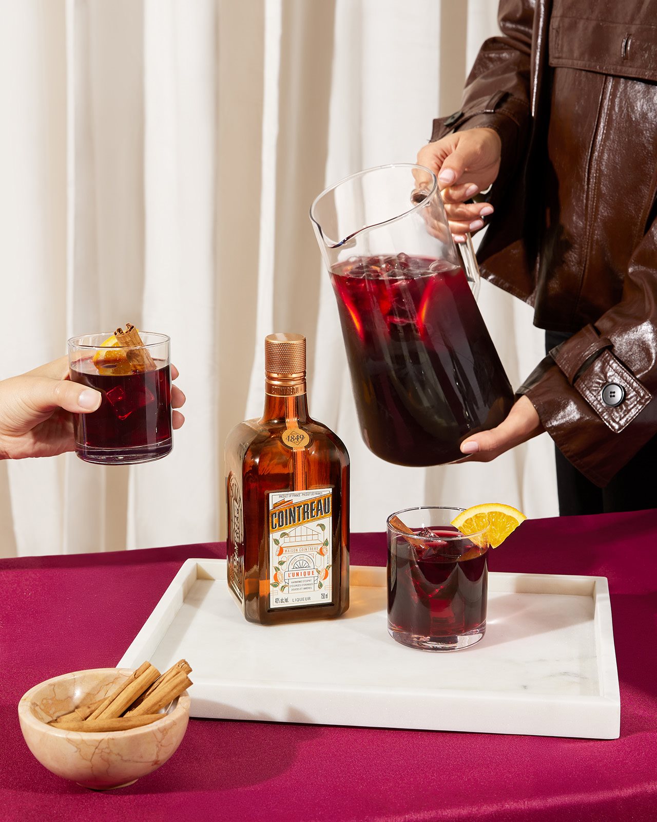 A woman serves a Winter Sangria in a pitcher to another person, with a bottle of Cointreau in the foreground.