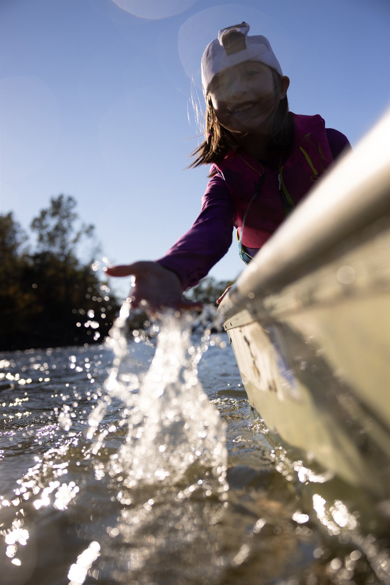 Little girl, wearing her cap backwards, using her hand to spash water while sitting on the side of a boat in the lake.