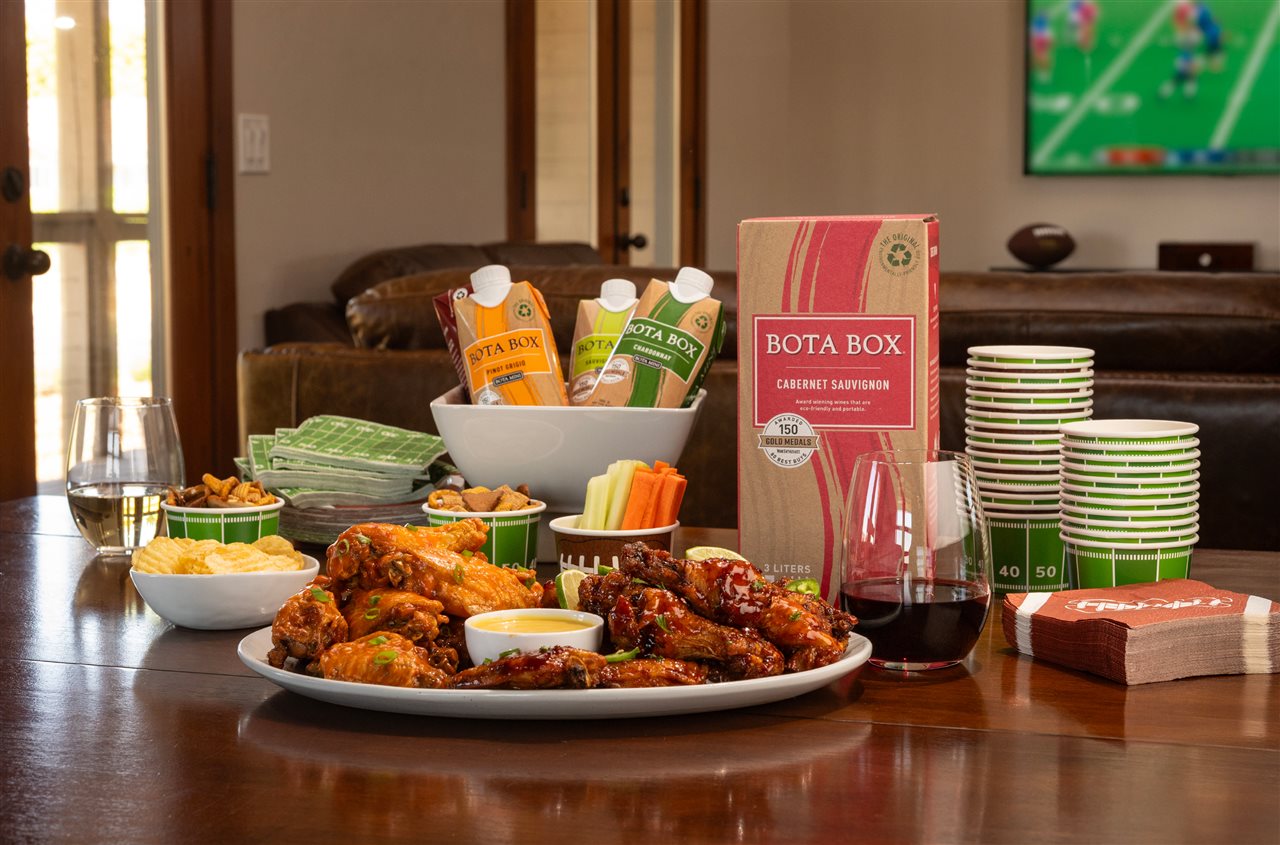 Table set with plateted chicken wings, cups, napkins and box of Bota Box wine.