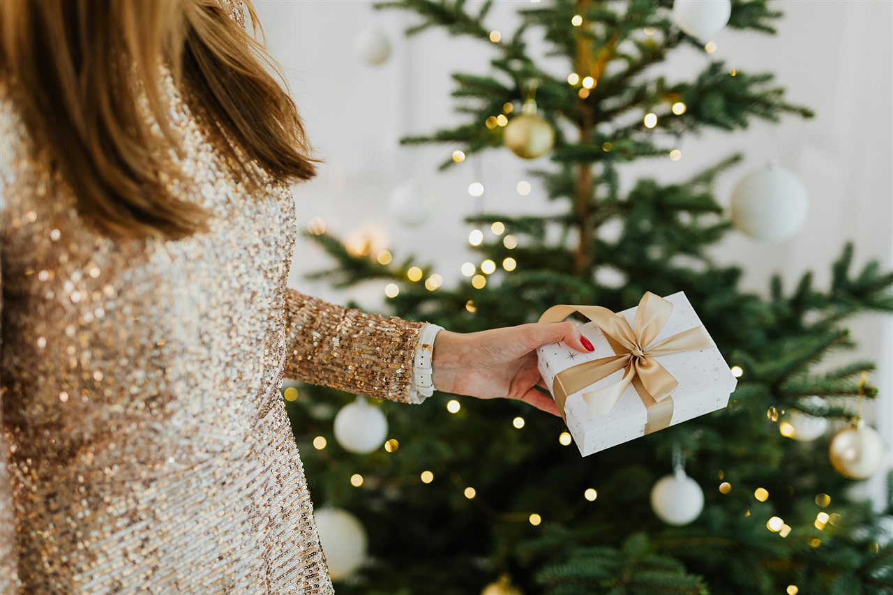 Woman wearing a sparkly dress holding a white package with gold ribbon and standing in front of a tree with white lights.