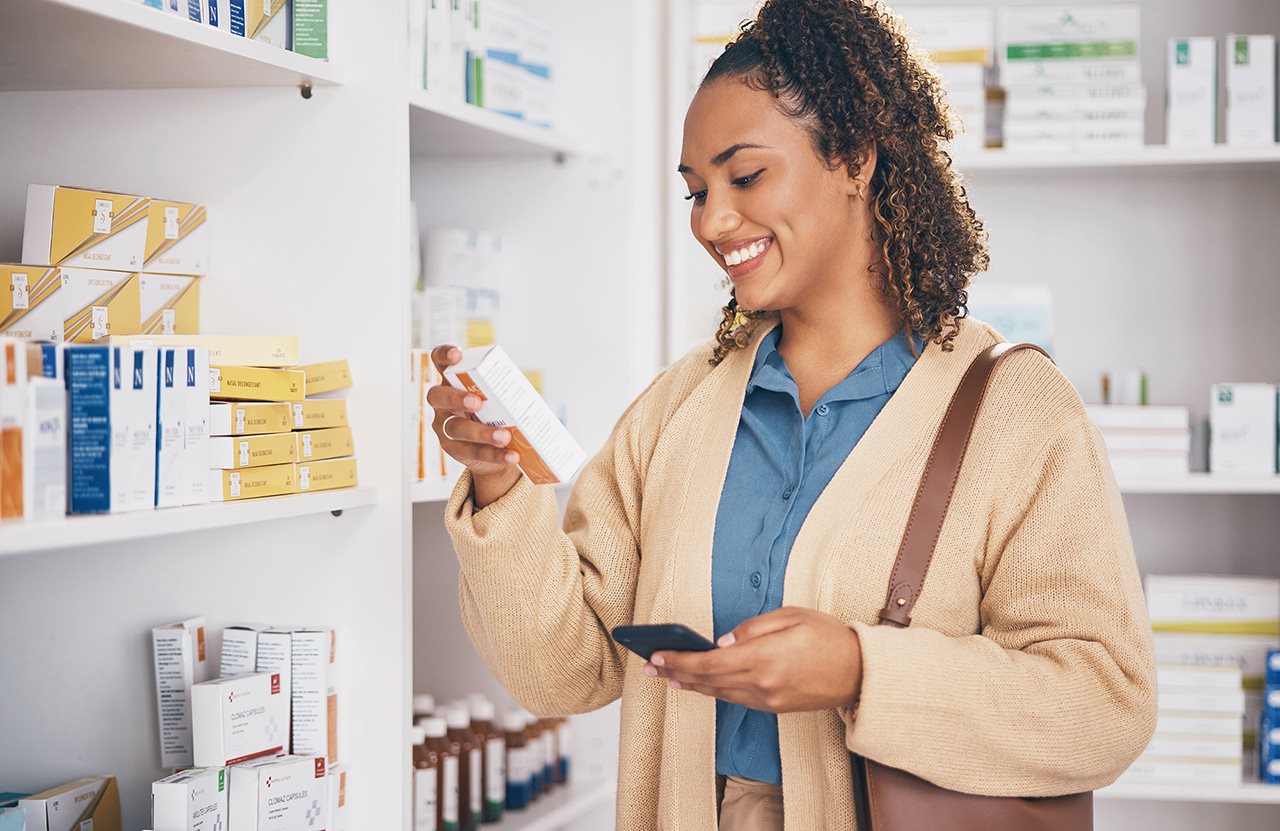 Smiling woman in a drug store reading the back of an OTC product while holding her phone.