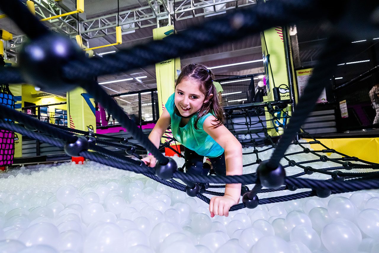 Girl on warrior ropes over ball pool.