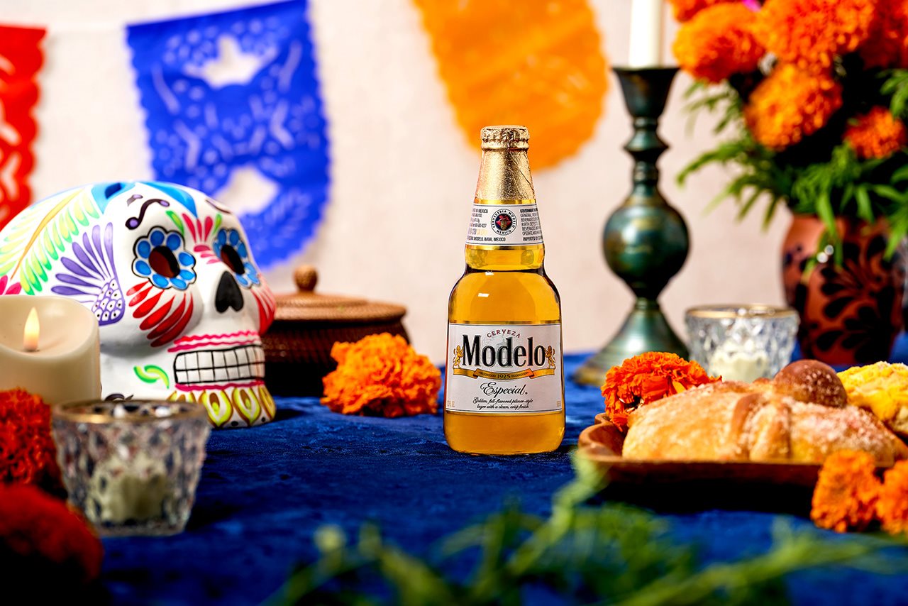 Primer plano de una botella de Modelo, una máscara del Día de los Muertos, velas y comida en un altar.
