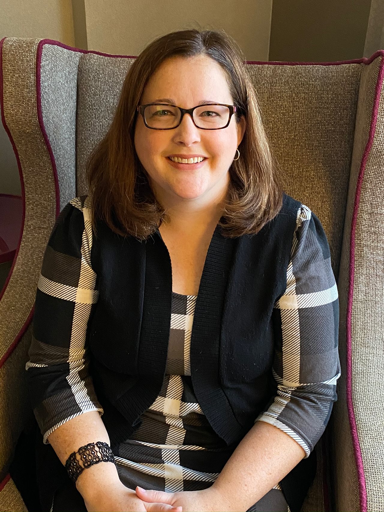 Smiling Alison wearing glasses and a plaid shirt and sitting in a wing-backed chair in the living room.