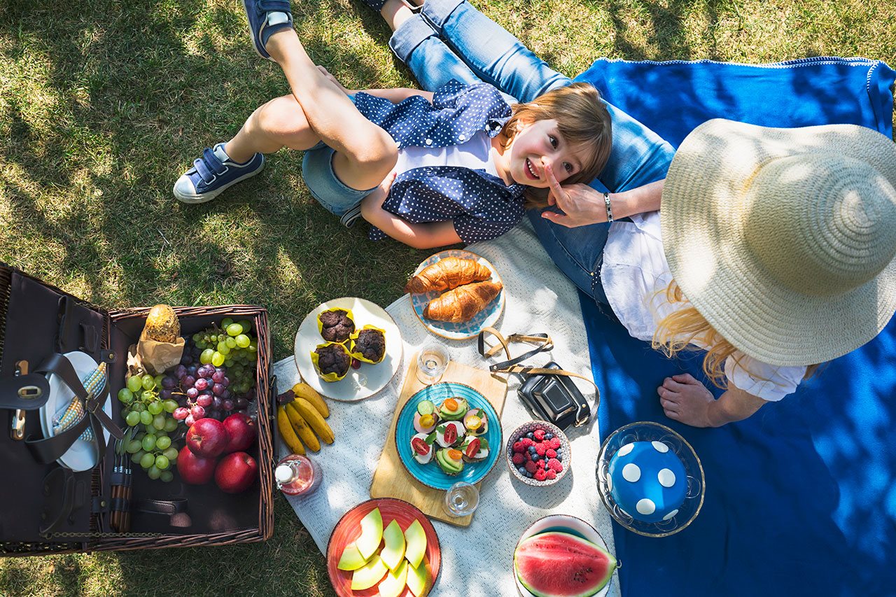 Mom and young daugher sitting on a blanket while enjoying a picnic under the shade of a tree.