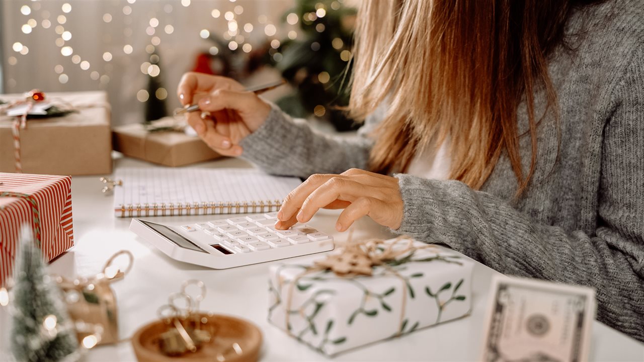 Woman checking over her holiday gift list with a calculator while sitting at a table at home surrounded by wrapped packages and holiday lights.