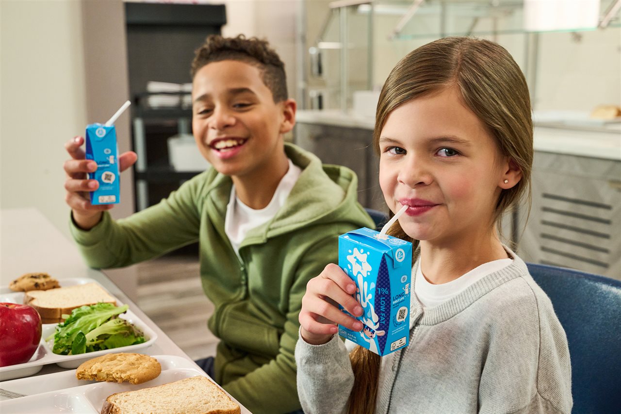 Two children sipping milk from a carton in a school cafeteria.