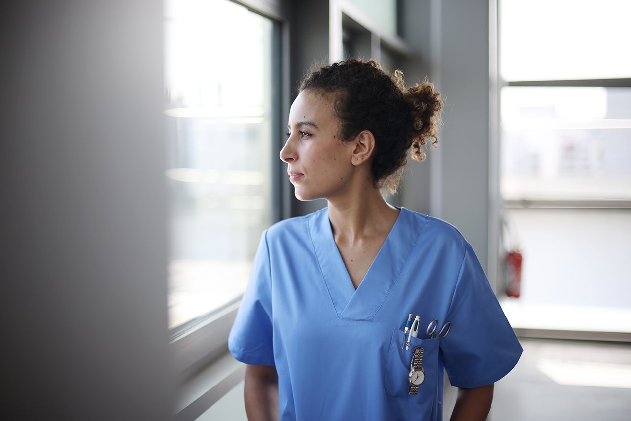 Nurse in blue scrubs in a hospital hallway looking out the window
