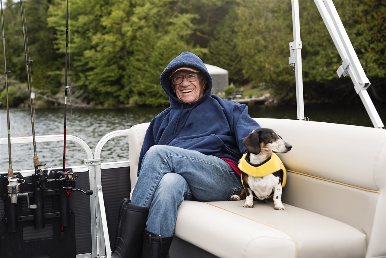 Senior man wears a coat and dog wears a life jackey while on a boat.